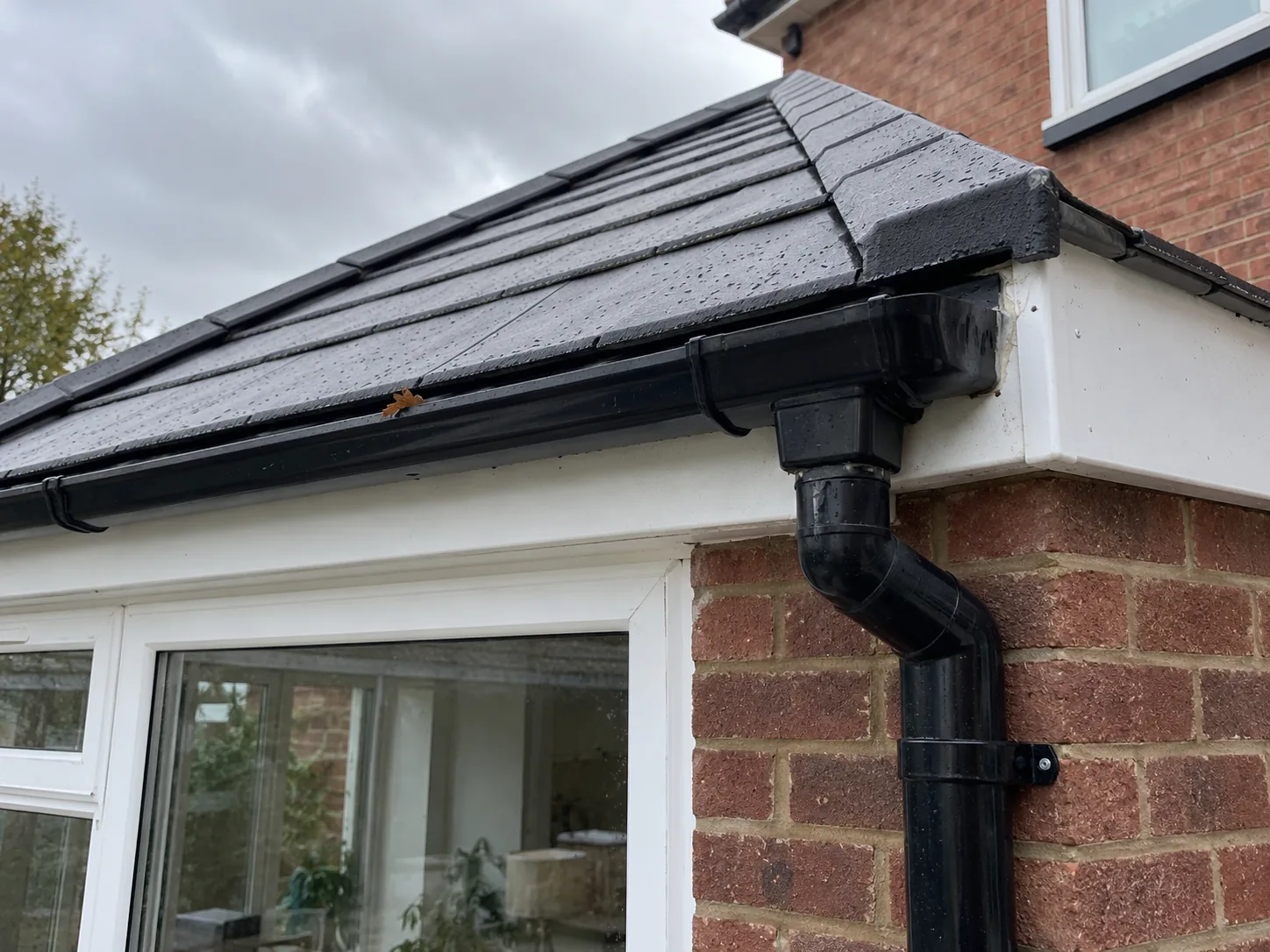 Close view of a new tiled conservatory roof edge in dark grey, wet from rain, with a white fascia, black gutter and downpipe against red brick