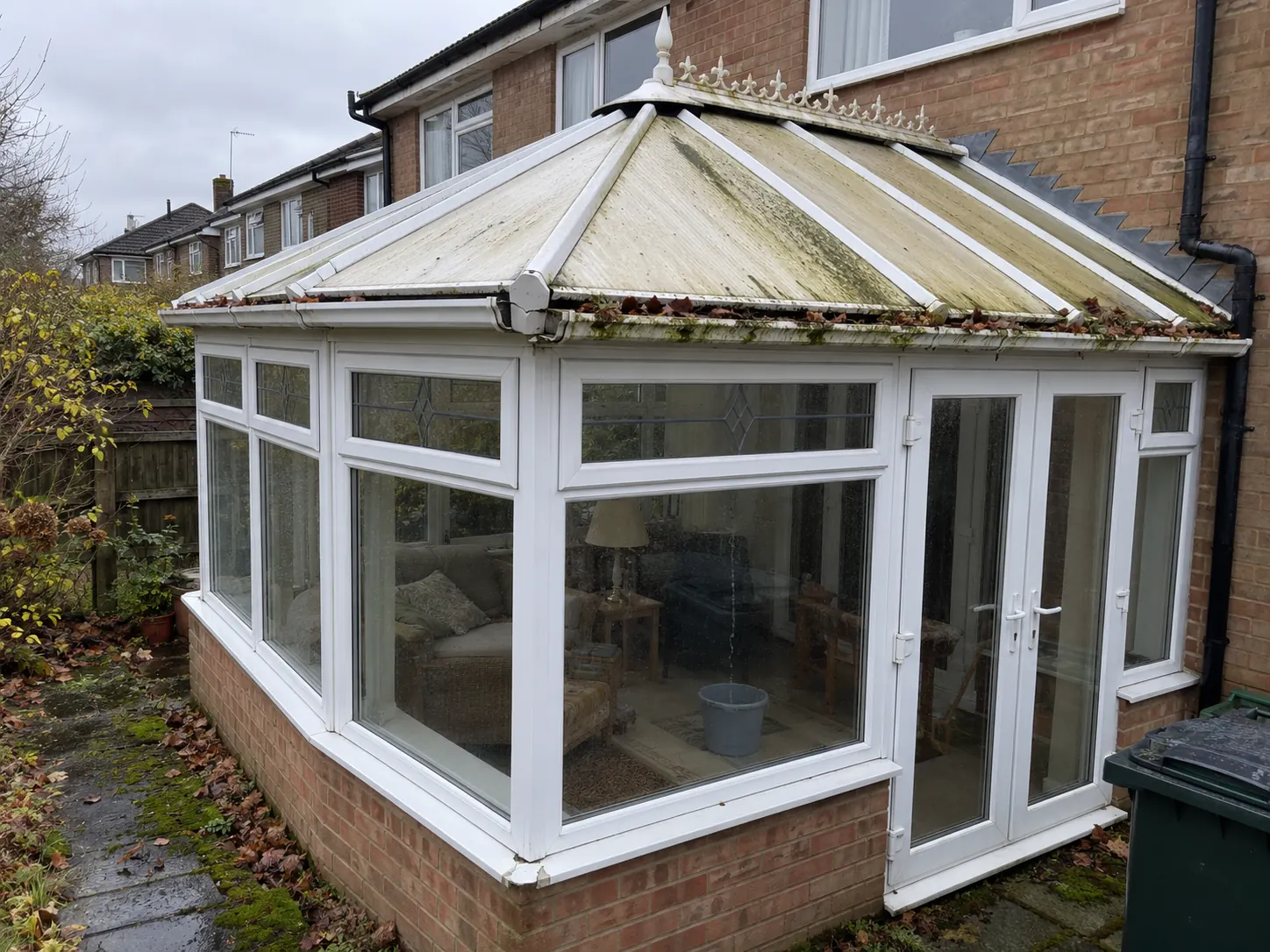 A tired white conservatory on a brick semi in grey drizzle, its polycarbonate roof clouded and streaked green, leaves packed in the gutter and a bucket on the floor inside